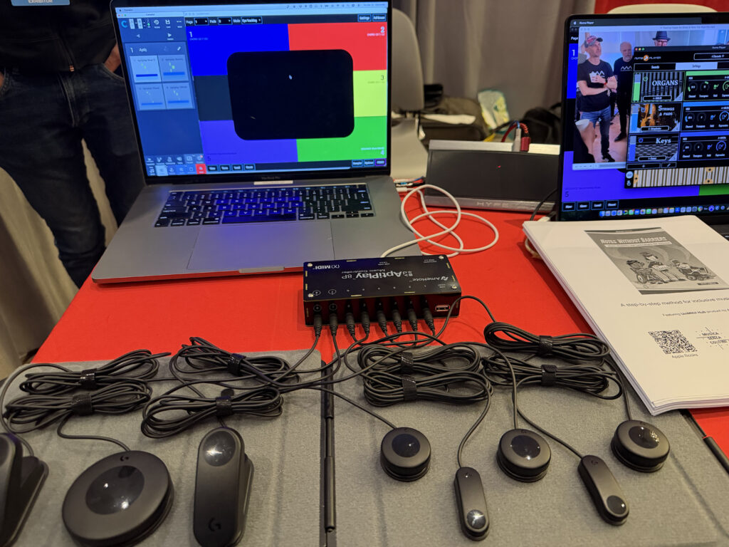 A table displays laptops with colorful screens, a SharpSpy device, several connected sensors with wires, and a document. Two people are visible on one laptop screen in a smaller video feed.