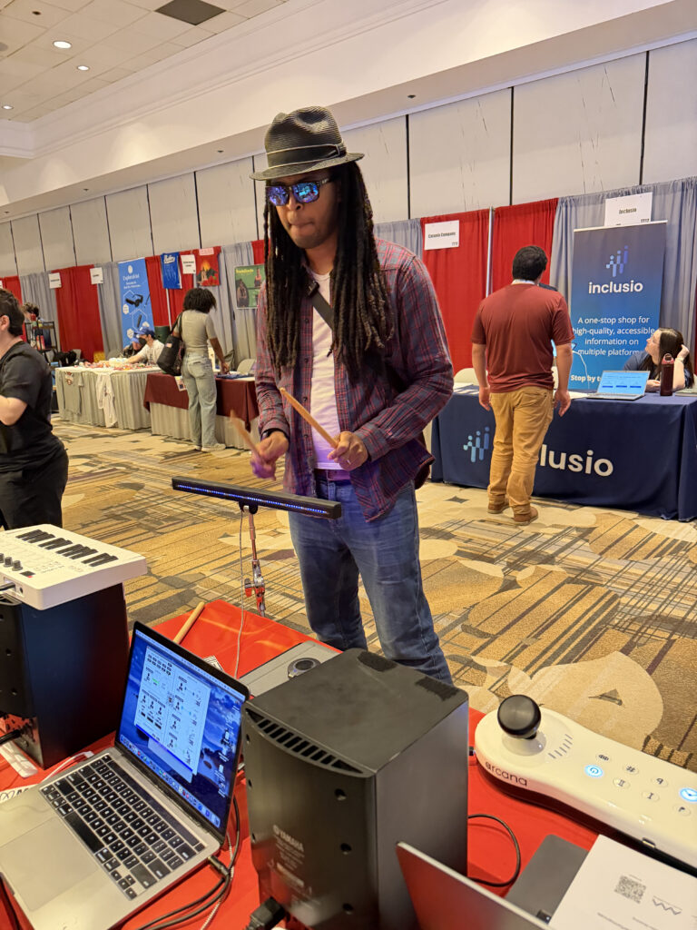 A man with long dreadlocks, sunglasses, and a fedora plays a theremin at a technology expo booth. Laptops and audio equipment are on the table in front of him. People and banners are visible in the background.