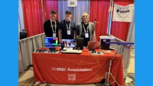 Lele Parravinic fro, Audio Modeling, Mike Ketn from Amenote and Athan Billias from MIDI2Marketing stand behind a red table displaying electronic music equipment at a booth for The MIDI Association. The table has the MIDI Association logo, and banners and gear are set up in the background.