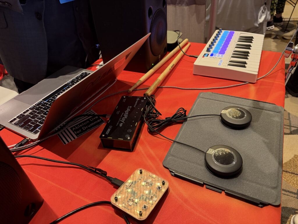 A close-up of an electronic music setup on a red table, featuring a laptop, MIDI keyboard, audio interface, two circular drum pads on a tablet, drumsticks, and various cables connecting the devices.