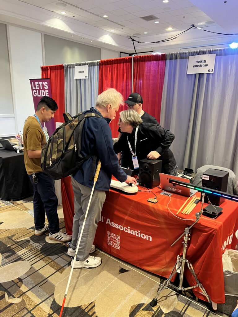 Two people assist a man with a white cane and backpack at an exhibition booth with a red tablecloth reading Association. Another person stands nearby. The booth has banners, tech equipment, and informational materials.