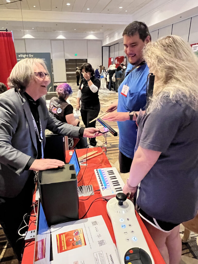 A smiling exhibitor in a gray blazer demonstrates an accessible music device to two attendees at a busy conference booth, featuring adaptive technology and informational displays on the table.