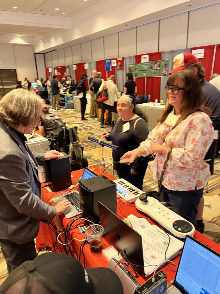 People interact with electronic music devices at a conference booth. A man demonstrates equipment on a table, while two women observe and engage, surrounded by other attendees and exhibition booths in a large hall.