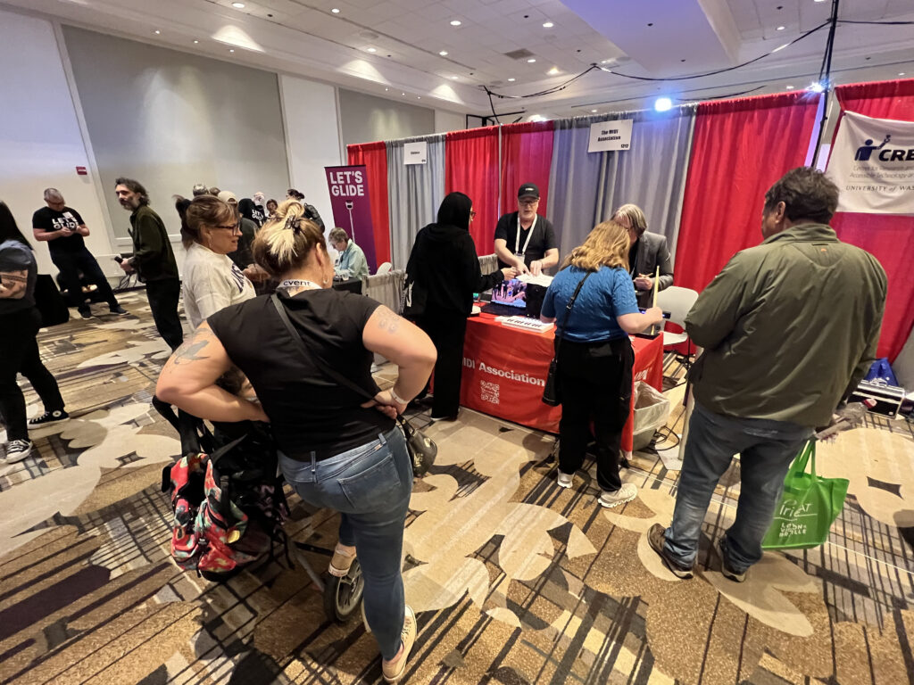 A group of people gather around a red table at an indoor convention booth, interacting with staff and materials. Red curtains and banners are visible in the background, along with informational displays.