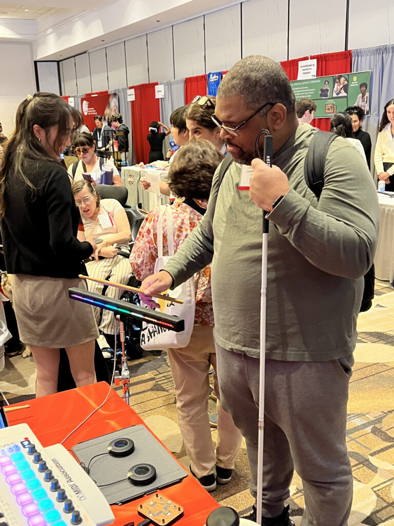 A man holding a white cane uses a tactile display at an exhibit booth, surrounded by people at a busy conference with informational tables and red and white curtains in the background.