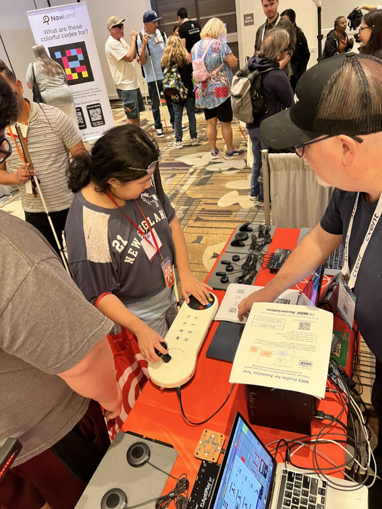 A young woman tries an accessible video game controller at an indoor event, while a man assists her. People walk and interact in the background near booths and informational posters.