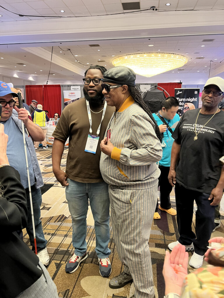 Two men pose together in a busy convention hall. One wears a brown shirt and badge; the other wears sunglasses, a striped suit, and a cap. Other people and booths are visible in the background.