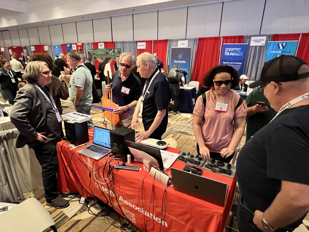 A group of people gather around a red table at a convention or expo, interacting with electronic equipment and laptops. Booths and banners are visible in the background, and attendees are engaged in conversation.