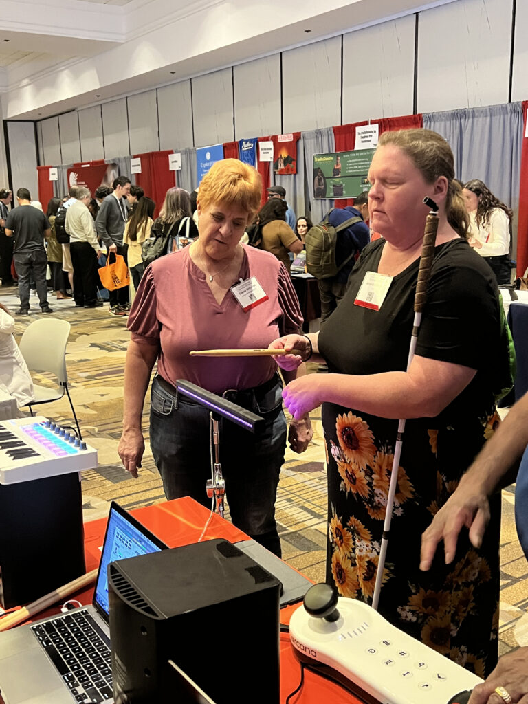 Two women stand at a booth with music equipment at an expo. One woman holds a white cane and uses a drumstick on an electronic instrument, while the other observes. People and display booths are visible in the background.