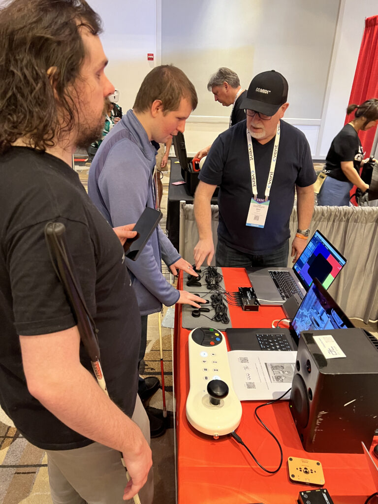 Three people stand at a booth with audio equipment and a computer; one man points at the laptop screen while others observe. The table has speakers, a large accessible controller, and informational materials.