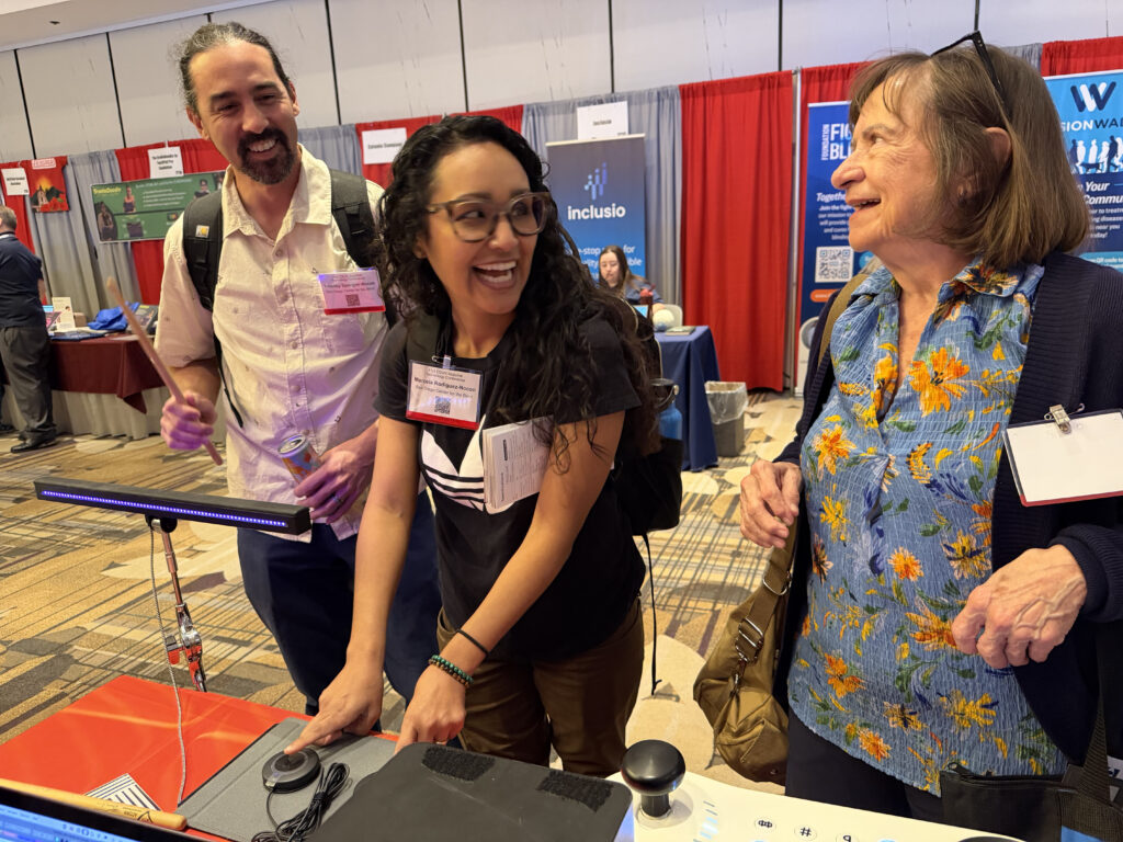 Three people smile and interact at a conference booth with a joystick control panel, surrounded by display booths and red curtains in the background.