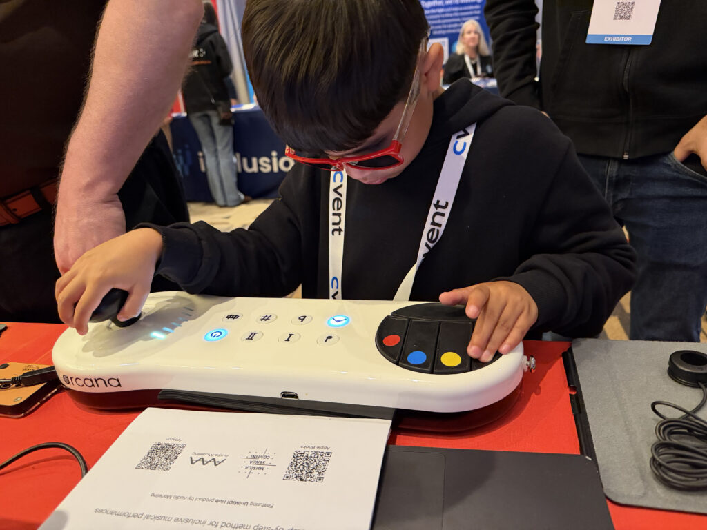 A young boy wearing dark glasses uses an accessible electronic device with large buttons and a joystick at an event booth. A printed page with QR codes and text is on the table in front of him.