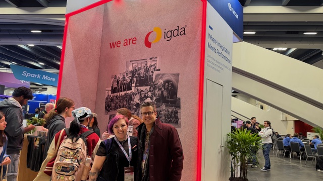 Two people stand smiling in front of an IGDA booth at an event. The booth features a collage of group photos, a sign reading “we are igda,” and several attendees walking around in the background.