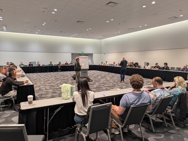 A large conference room with people seated around a U-shaped table layout, attentively listening to two speakers standing in the center near a flip chart. The room is well-lit with a modern design.