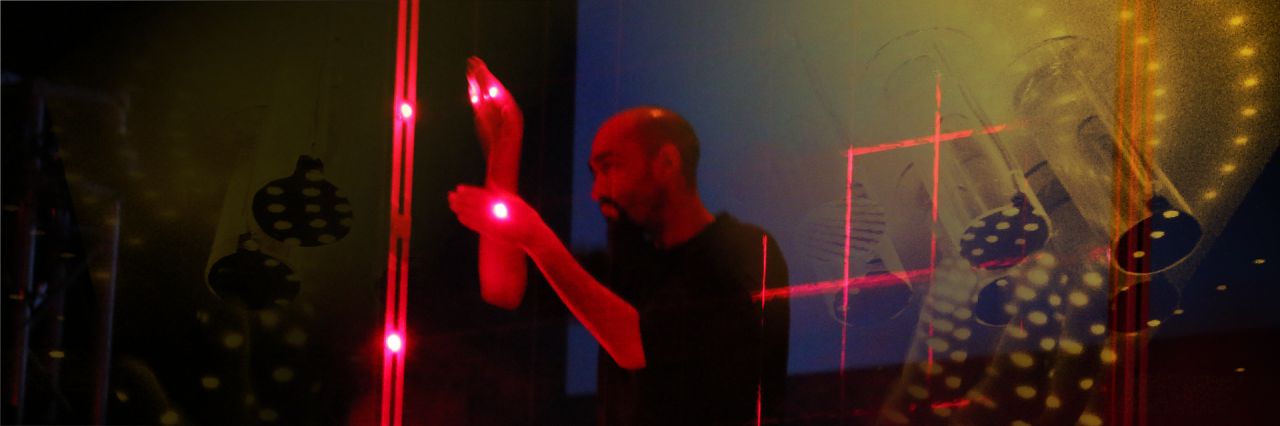 A person interacts with red lasers or light beams in a dark room, with reflections and abstract light patterns on transparent surfaces around them.