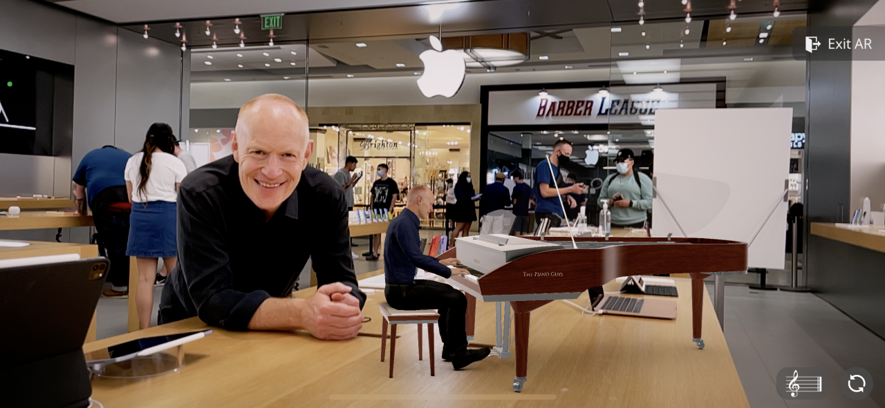 A smiling man leans on a wooden table in an Apple Store, while a miniature pianist sits at a grand piano on the same table. Shoppers and staff are seen in the background near a glass entrance.