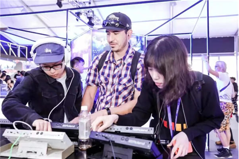 Three people interact with electronic music equipment at an indoor event. Two are focused on using the keyboards, while the third stands behind them observing. The scene is lively, with other attendees in the background.