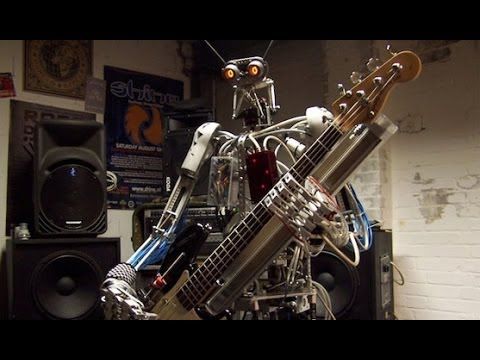A humanoid robot with mechanical arms is holding an electric guitar in a room filled with speakers and posters on the brick walls.