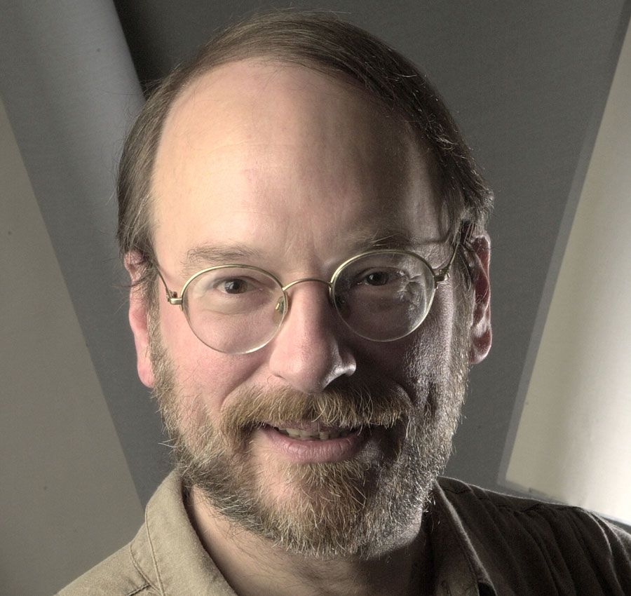 A middle-aged man with glasses, a beard, and mustache smiles gently. He has thinning light brown hair and is wearing a beige shirt. The background is softly lit with geometric shapes.