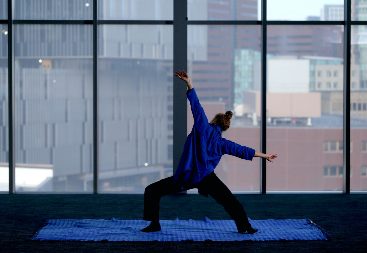 A person practices a yoga pose on a blue mat indoors, facing large windows with a cityscape view of modern buildings in the background.
