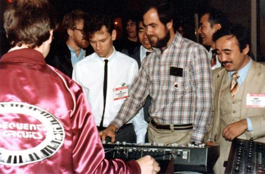A group of men, some with badges, gather around a synthesizer or music equipment. One man wears a red jacket with Sequential Circuits on the back. The mood appears focused and engaged.