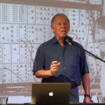 A man stands at a podium with a laptop and microphone, speaking. Behind him is a projected black-and-white image of a younger man and a large vintage synthesizer with many knobs and cables.
