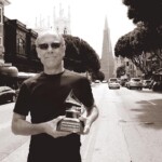 A man wearing sunglasses and a black shirt stands in the middle of a city street, smiling and holding a Grammy award. Tall buildings and cars line the street, with the Transamerica Pyramid visible in the background.