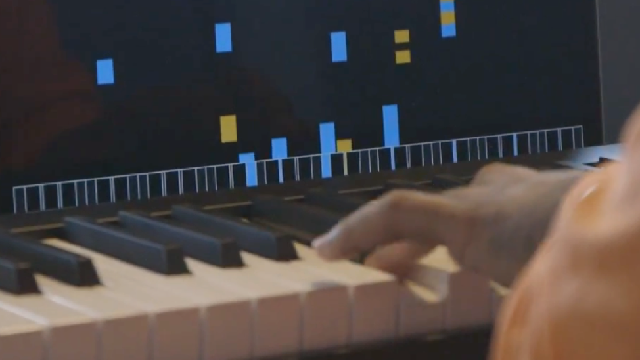 A person plays a piano keyboard while a screen displays a digital piano roll with blue and yellow notes indicating music being played.