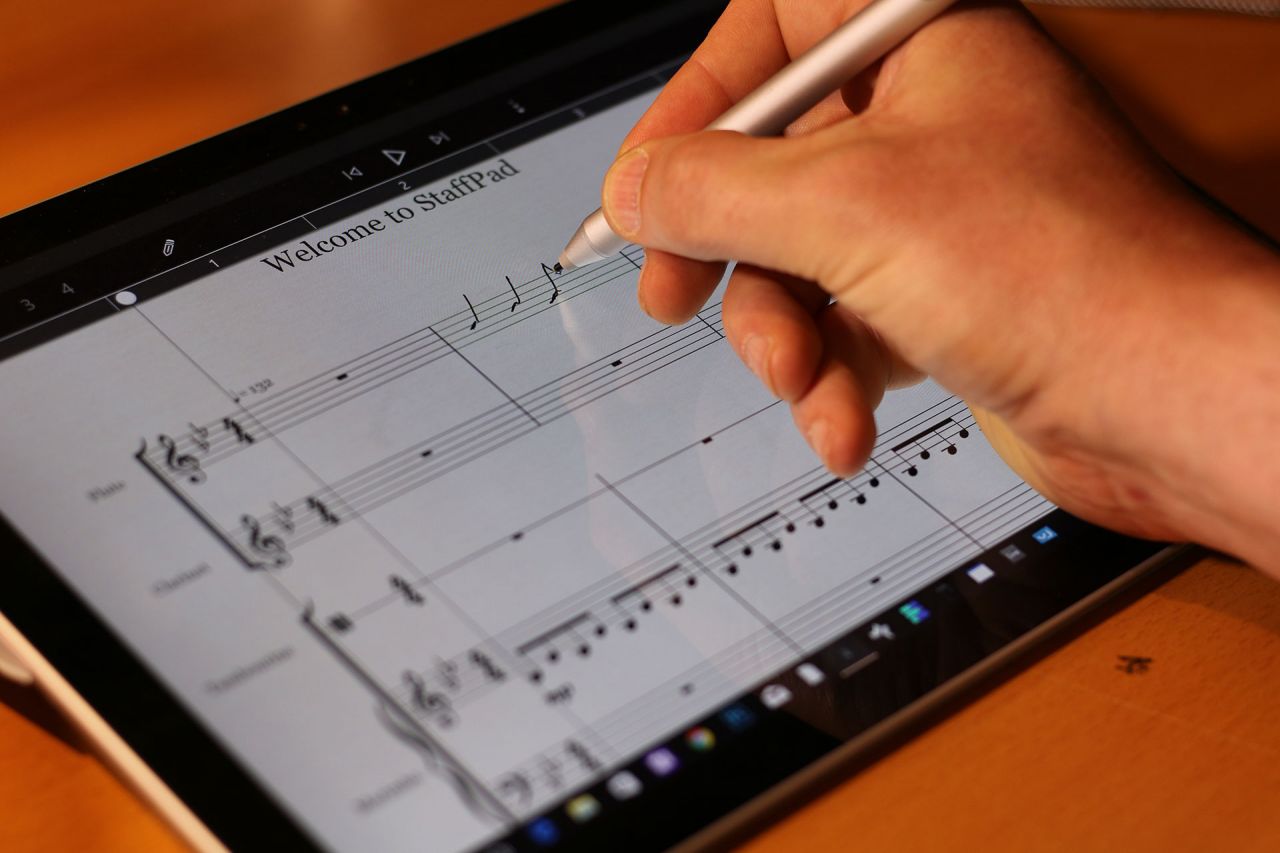 A hand uses a stylus to write or edit sheet music on a tablet screen displaying music notation software. The music score on the screen is titled Welcome to StaffPad.