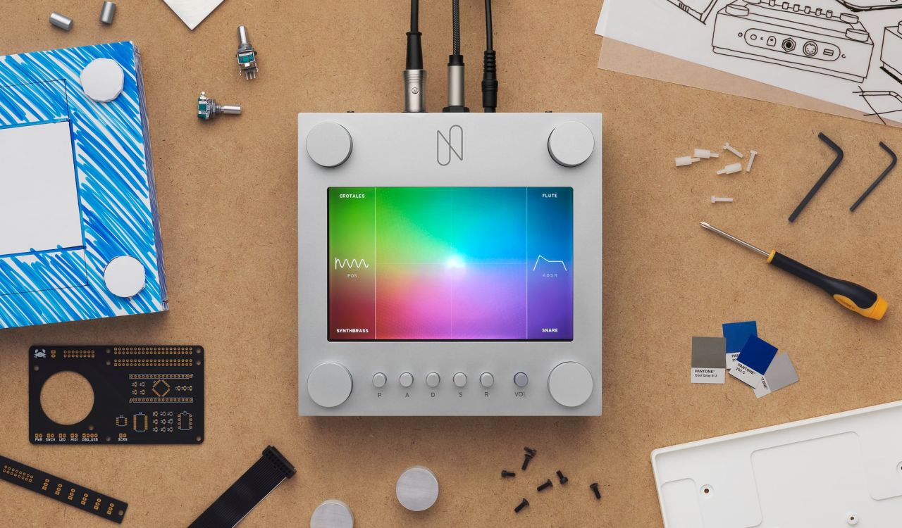 A modern electronic music device with a colorful touchscreen and four knobs sits on a workbench surrounded by tools, electronic components, sketches, and color swatches.